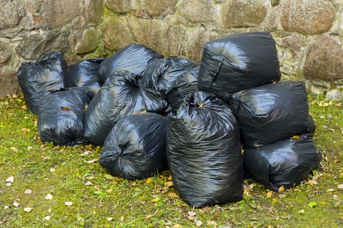 A collection of nine black plastic rubbish bags, tightly sealed and piled on top of each other in front of a weathered stone wall, situated on a grassy area with scattered fallen leaves. The bags show a glossy surface reflecting natural light, indicating they are filled with waste material. The surrounding environment suggests an outdoor setting, possibly a residential or commercial outdoor space, with the bags likely awaiting collection or disposal. The background features a stone wall composed of irregularly shaped rocks with visible moss and weathering, contributing to an aged appearance. The scene is evenly lit, highlighting the texture of the bags, grass, and stone wall. The presence of the bags aligns with rubbish removal services provided by Office Clearance Chingford, serving the Chingford area nearby.