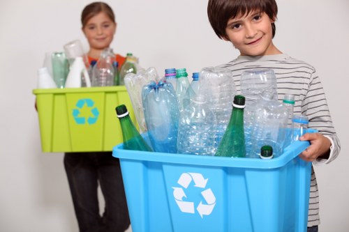 A young boy standing in front of a white background is holding a bright blue recycling bin filled with clear plastic bottles, some with green caps, all placed upright and tightly packed. Slightly behind him, a young girl is nearby holding a lime green recycling bin that contains a similar assortment of plastic bottles and containers, also upright, and features a blue recycling symbol on the side. The plastic bottles in both bins include water bottles and beverage bottles made of transparent and slightly textured plastic, with some showing ridges or embossed patterns. The scene emphasizes the act of recycling, relating to rubbish removal services, with the children demonstrating proper sorting of recyclable plastic waste, suitable for local environmental management practices. The vibrant colours of the bins contrast against the plain background, highlighting the importance of waste segregation in eco-conscious disposal efforts typical to Chingford and the surrounding London area. The image captures the concept of environmental responsibility in domestic rubbish recycling, aligning naturally with rubbish removal services offered by Office Clearance Chingford.