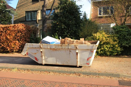 A large, white metal skip positioned on a paved residential street in Chingford, containing a mixture of cardboard boxes and packing materials. The skip shows signs of rust and weathering, with reddish-brown stains along its edges and surface. It is situated immediately adjacent to a sidewalk with a low brick curb, in front of a tidy garden with well-maintained bushes and trees, some with autumnal foliage. In the background, there are two detached houses with brick facades, large windows, and sloped tiled roofs, partially obscured by trees and shrubs. The scene is illuminated by natural daylight, typical of a clear day, contributing to a professional, observational portrayal suitable for a rubbish removal service in the local area, such as office clearance in Chingford as offered by Office Clearance Chingford.