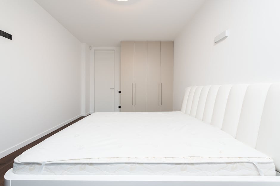 In an outdoor urban setting with graffiti-covered concrete walls serving as a backdrop, two makeshift beds are placed directly on the concrete ground. The first bed consists of a flat, rectangular foa